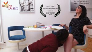 Oral Exam, a film by Ms. Naughty. A man knees in front of a woman who looks gleeful, study items are on the table nearby. Sexiest International Film, Toronto International Porn Festival 2017